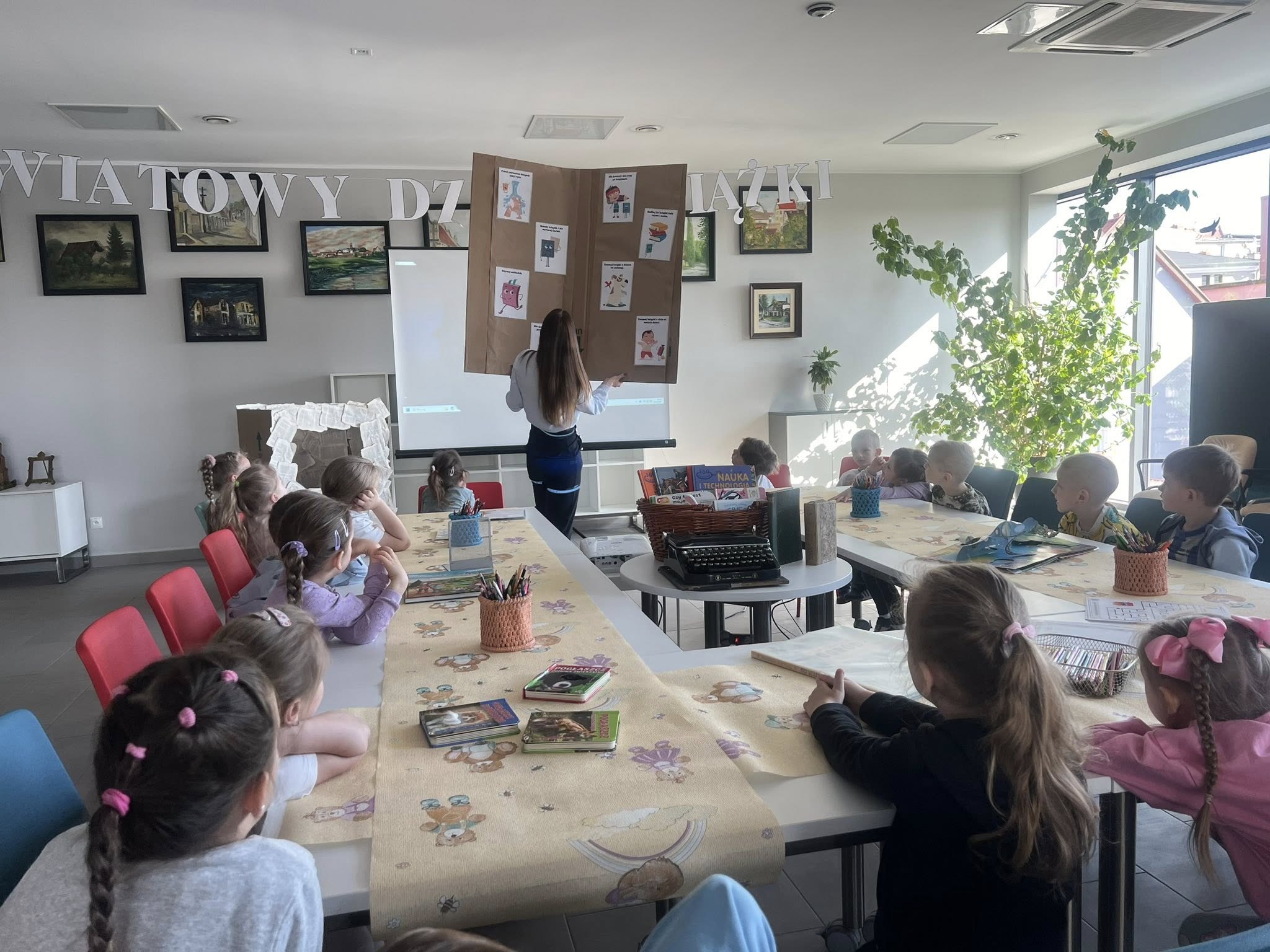 Dzieci zgromadzone wokół stołu. Bibliotekarka prezentuje dużą kartonową książkę.