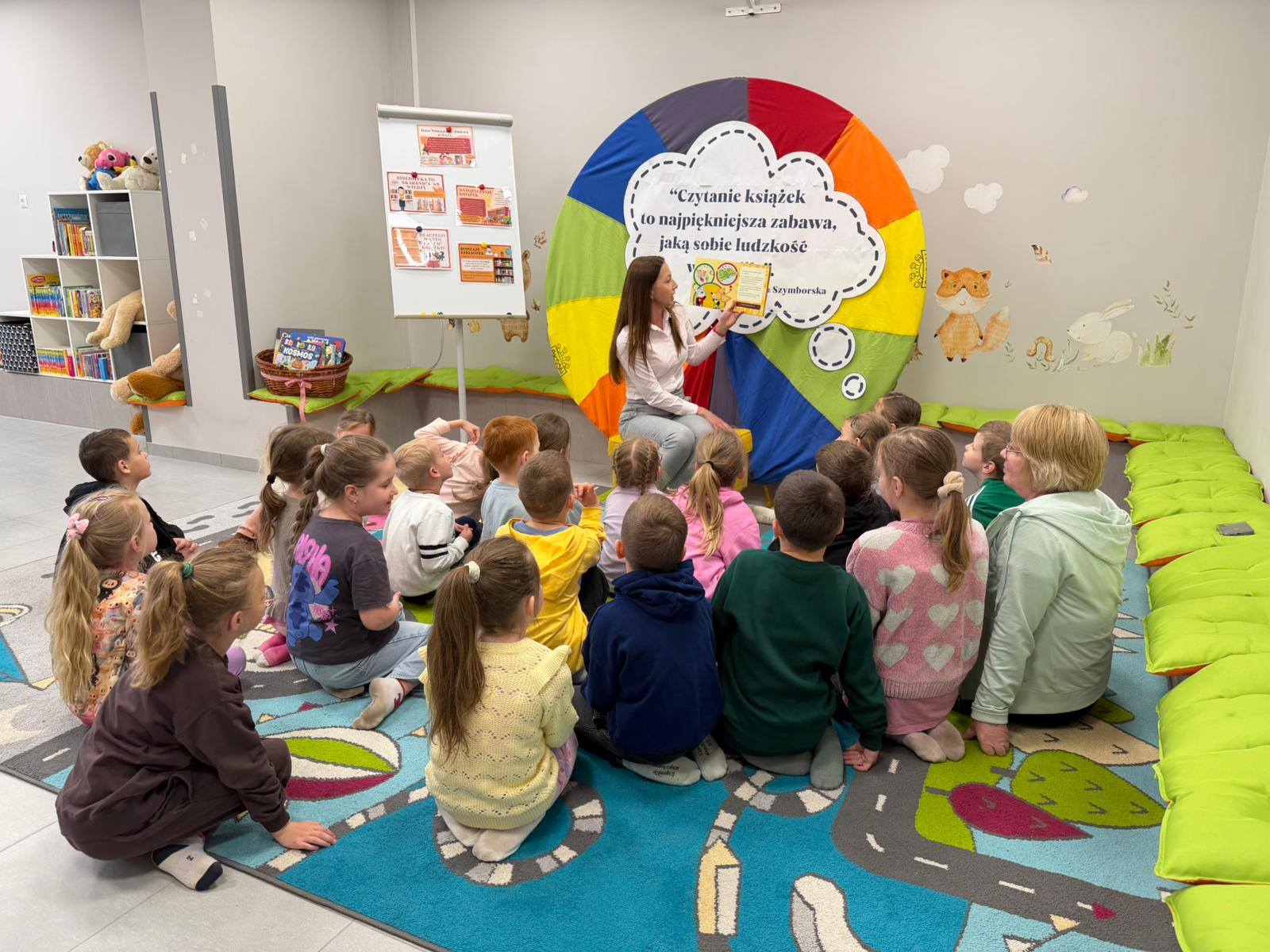 Przedszkolaki siedzą przed bibliotekarką na dywanie i oglądają prezentowaną książkę.