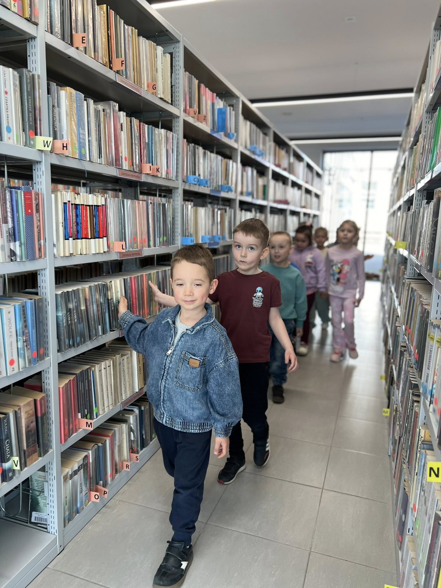 Dzieci zwiedzają bibliotekę, przechodząc między regałami.