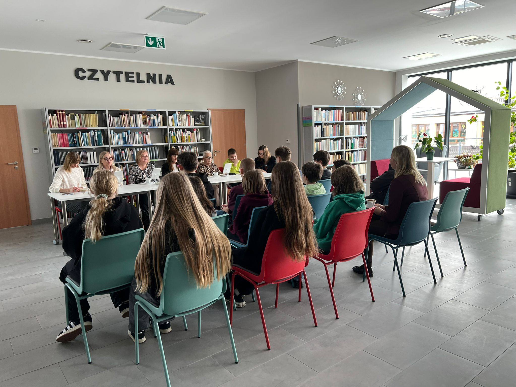 Grupa dzieci z klasy 6 Szkoły Podstawowej w Margoninie oraz nauczycielka siedzą na krzesłach jako widownia i wysłuchują czytających, którzy siedzą za stołem. Wśród czytających jeden uczeń.