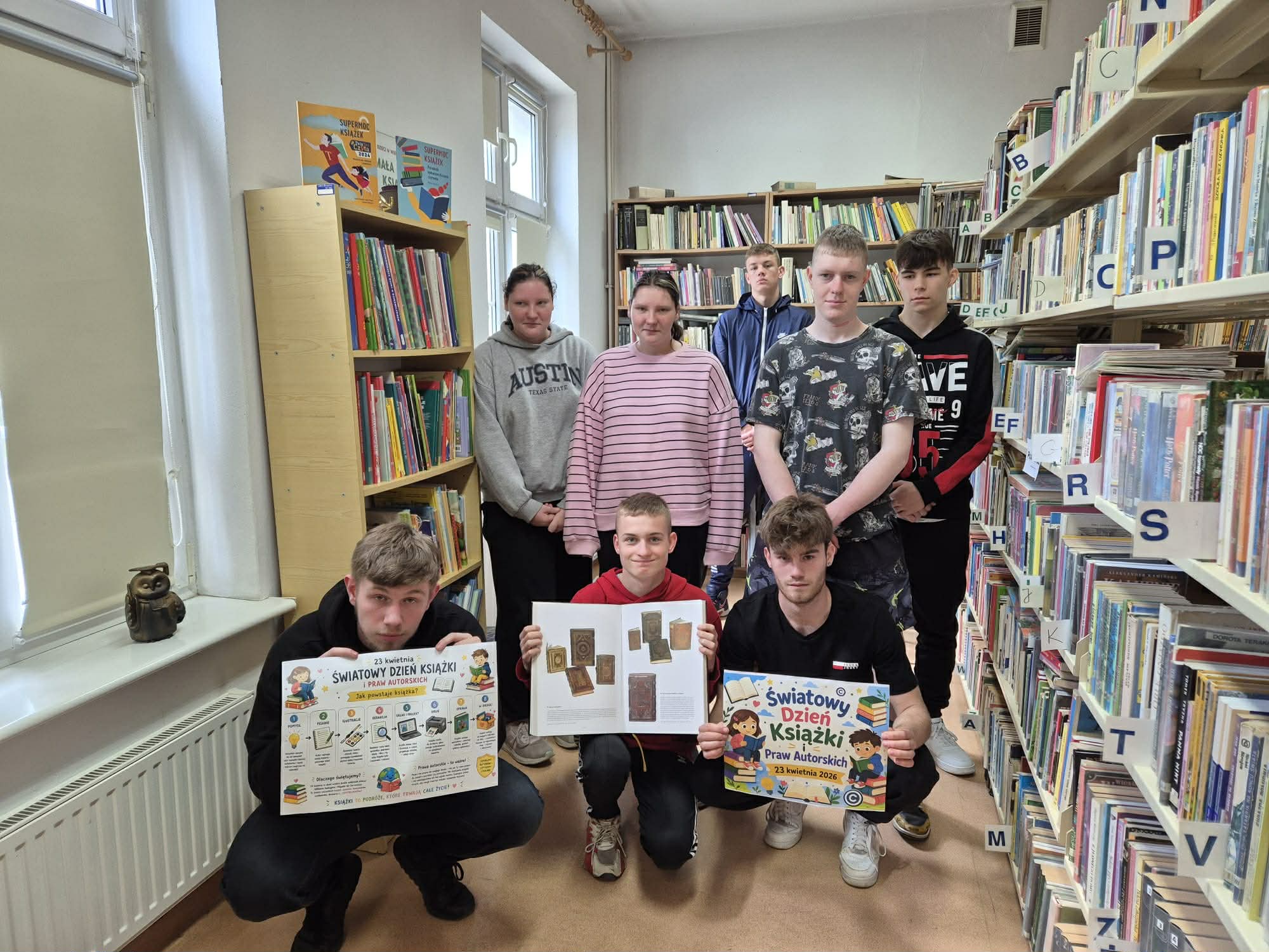 Uczniowie ze szkoły ponadpodstawowej prezentują otwarte książki stojąc na tle regałów w bibliotece.