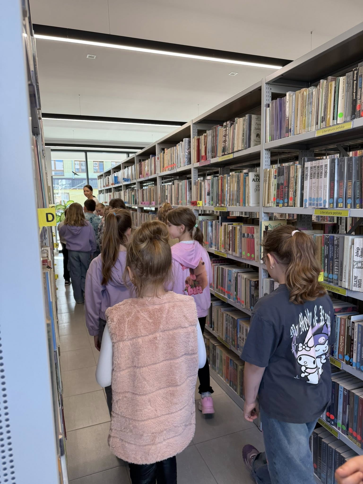 Drugoklasiści zwiedzają bibliotekę spacerując między regałami.