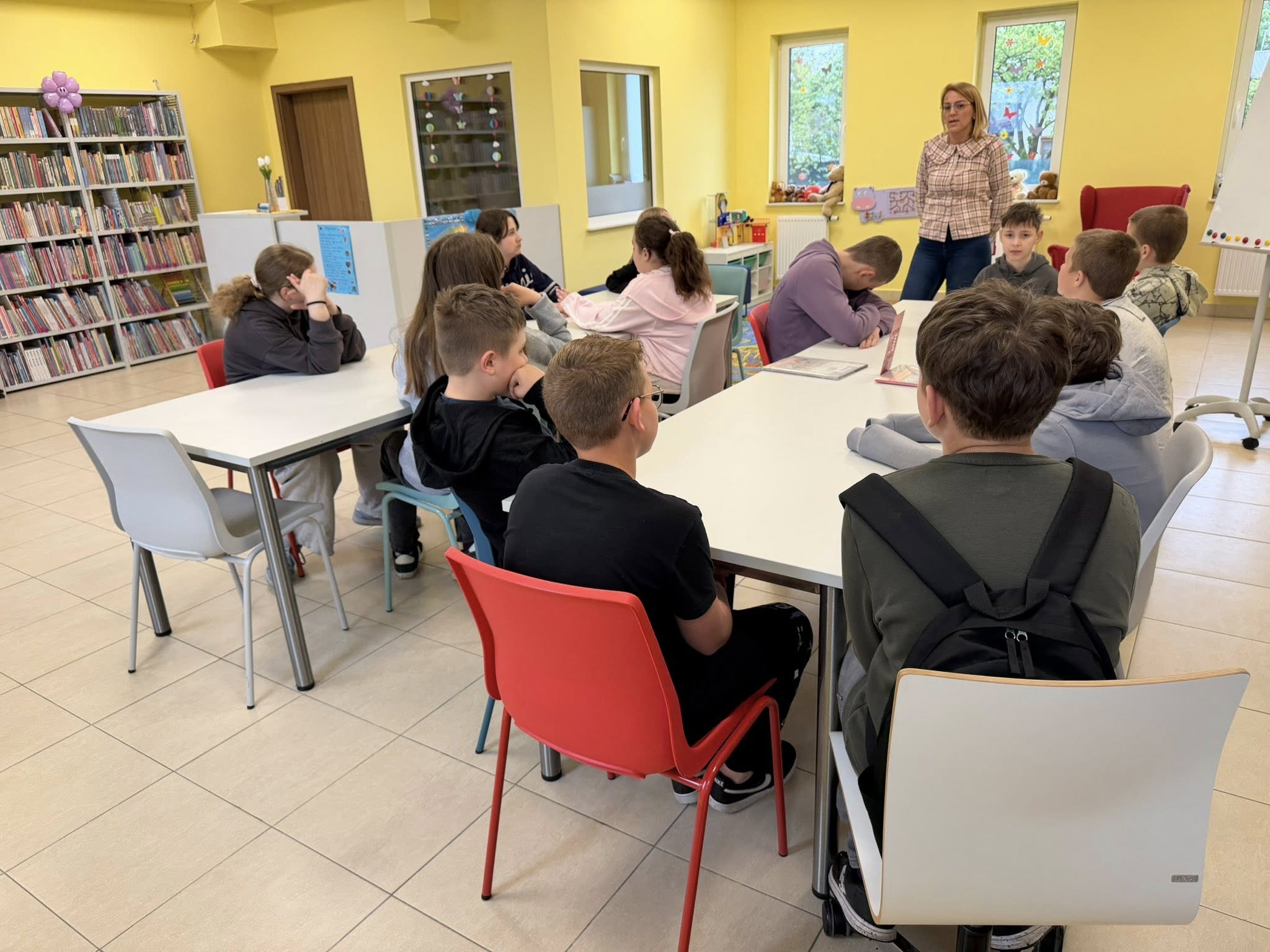 Szóstoklasiści siedzą przy stołach, bibliotekarka stoi przed nimi i prowadzi zajęcia.