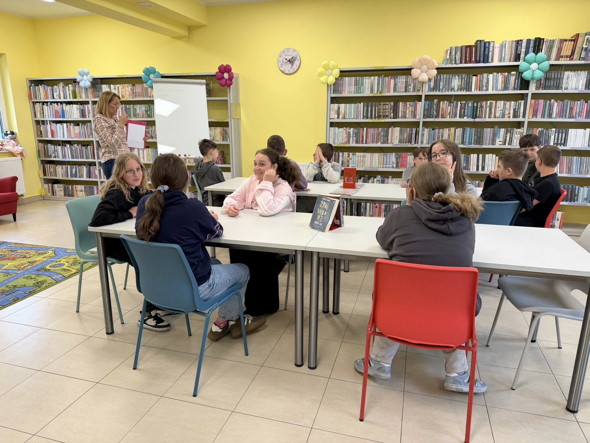 Szóstoklasiści siedzą przy stołach, bibliotekarka stoi przy tablicy magnetycznej i pokazuje trzymane w ręce materiały.