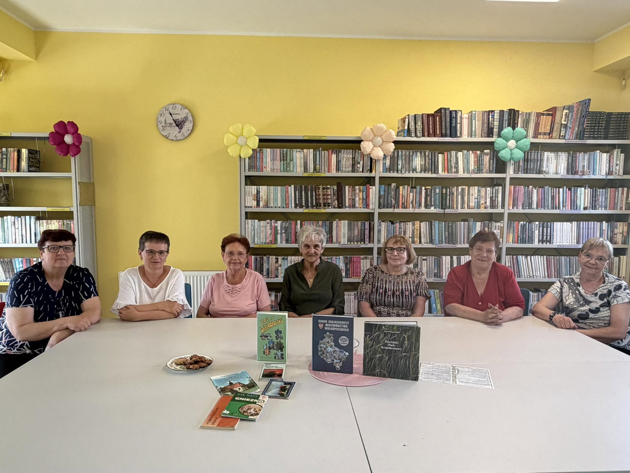 Siedem kobiet w wieku emerytalnym pozuje do zdjęcia siedząc przy stole. Na stole stoją książki.