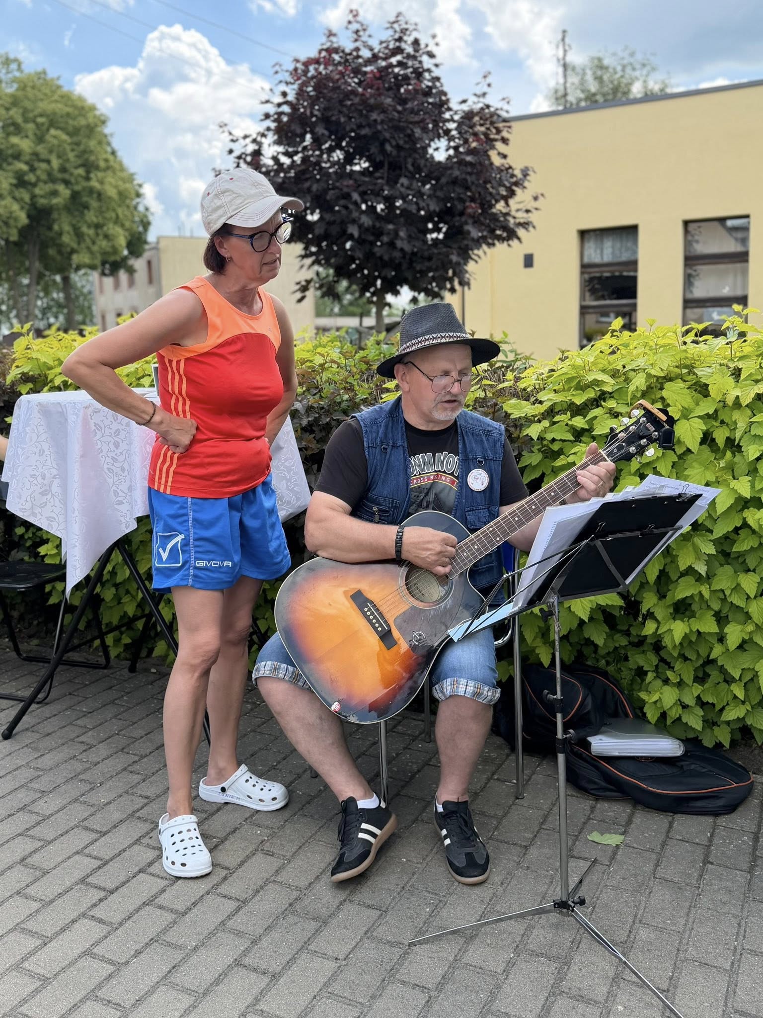Gitarzysta siedzi, kobieta stoi nad nim i patrzy w śpiewnik.