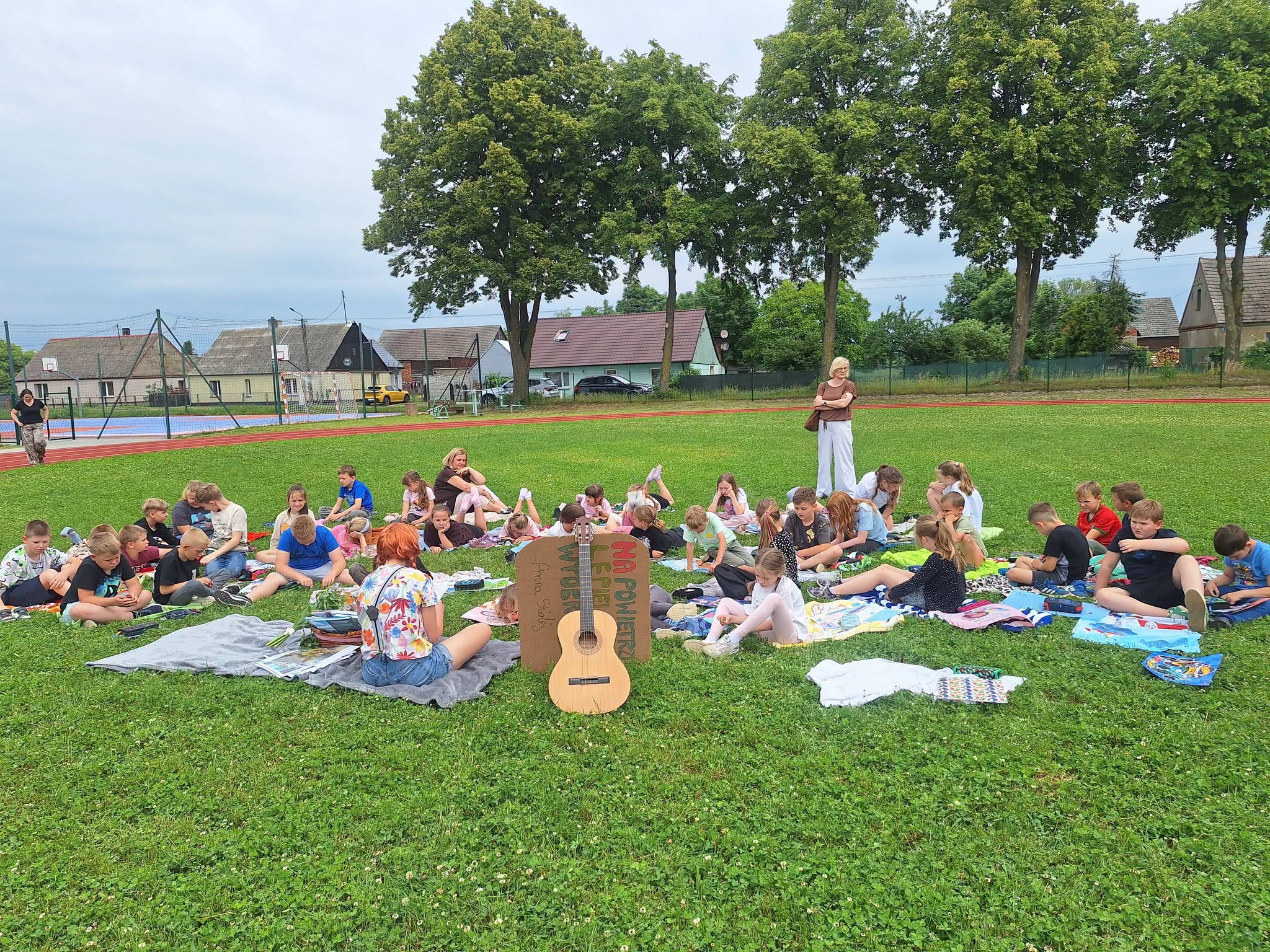 Grupa składająca się z uczniów i nauczycielki siedzi na kocu. Jedna nauczycielka stoi. Na pierwszym miejscu gitara.