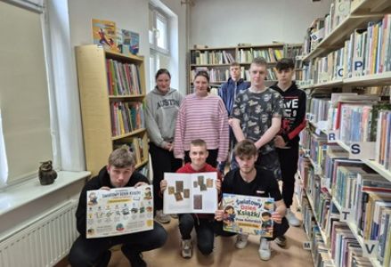 Uczniowie ze szkoły ponadpodstawowej prezentują otwarte książki stojąc na tle regałów w bibliotece.