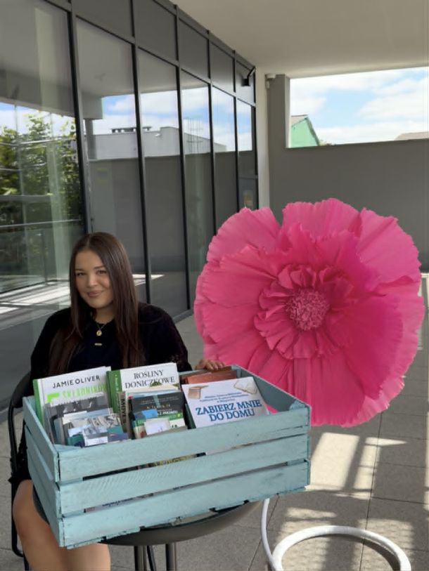 Bibliotekarka siedzi przy stoliku, na nim książki w koszu. Obok ozdobny kwiat.