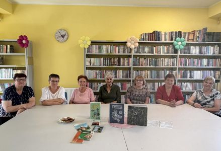 Siedem kobiet w wieku emerytalnym pozuje do zdjęcia siedząc przy stole. Na stole stoją książki.