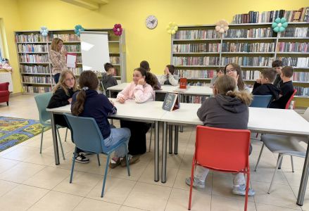 Szóstoklasiści siedzą przy stołach, bibliotekarka stoi przy tablicy magnetycznej i pokazuje trzymane w ręce materiały.